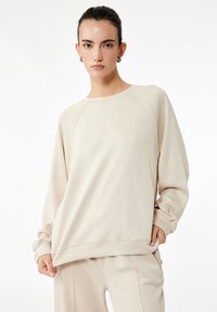 Sudadera de color beige claro con cuello redondo, mangas raglán y tela suave. Su corte relajado presenta un dobladillo que se superpone ligeramente.