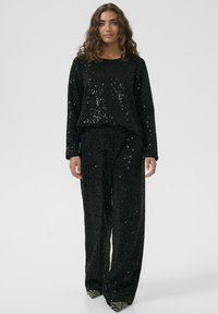 Černý souprava loungewear s flitry, která se skládá z volného dlouhého rukávu a širokých kalhot, obě zdobené třpytivou texturou.