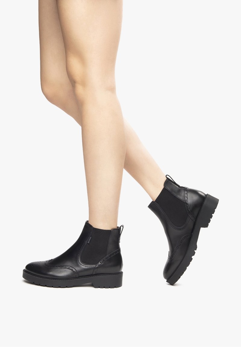 Jambes portant des bottines Chelsea en cuir noir avec des côtés élastiques et des détails décoratifs brogue sur fond blanc.