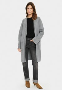 Cardigan grigio con maniche lunghe e apertura frontale, realizzato in tessuto morbido. Indossato sopra un top nero, abbinato a jeans scuri e tacchi marroni.