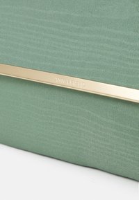 Surface en tissu vert avec une barre métallique horizontale dorée gravée du texte "ANNA FIELD" près du centre.