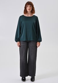 Blusa di raso color teal con maniche lunghe, abbinata a jeans scuri in denim a vita alta e gamba larga. Il modello indossa stivaletti neri alla caviglia.