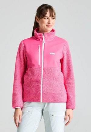 Una mujer luce una chaqueta polar de color rosa intenso, con cuello alto, y unos pantalones blancos sobre un fondo claro y liso.
