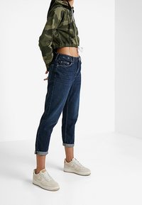 Maskáčová krátká mikina s kapucí a stahovací šňůrkou, tmavě modré zúžené džínové kalhoty a světle béžové tenisky s bílými detaily. Otočené manžety na džínách.