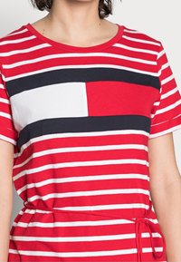 Punainen ja valkoinen raidallinen t-paita, jossa tummansiniset yksityiskohdat ja valkoinen neliömäinen pata; pehmeää puuvillakangasta ja rento malli, jossa on solmittu vyötäröyksityiskohta.