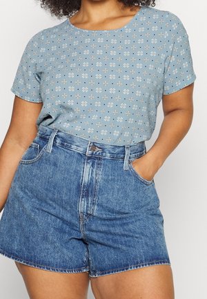 Vrouw draagt een lichtblauw bloemenprint kort mouw shirt, ingestopt in een hoge blauwe spijkershort, met één hand in de zak.