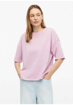 Kobieta o prostych włosach, w luźnym jasnofioletowym t-shircie i jasnoniebieskich dżinsach, z rękami w kieszeniach, patrząca na bok.