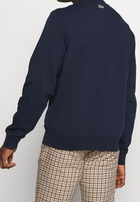 Námořnicky modrá mikina s kulatým výstřihem z měkké látky, s žebrovanými manžetami a lemem, s malým logem na zádech. Nošena s károvanými kalhotami.