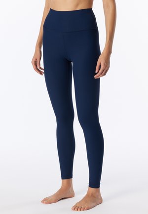 Frau trägt hoch taillierte, marineblaue gerippte Leggings und steht barfuß vor einem schlichten weißen Hintergrund.