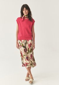 Haut sans manches en tricot rouge associé à une jupe midi beige à motifs floraux avec des feuilles vertes et des fleurs rouges. Le mannequin porte des sandales beige clair.