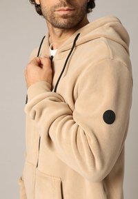 Hoodie en polaire beige avec une poche kangourou à l'avant, des cordons de serrage noirs, et un écusson rond noir sur la manche gauche. Texture douce.