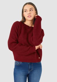 Donna che indossa un maglione bordeaux con piccoli ornamenti vicino al collo e jeans blu, posando con una mano vicino al viso.