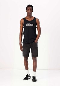 Svart ermeløs tanktop med "oblivion" grafikk, kombinert med mørk denimshorts. Brukt med svarte joggesko og hvite ankelsokker.