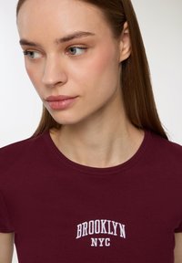 T-shirt a righe bordeaux con scritta bianca "BROOKLYN NYC", colletto rotondo e maniche corte. Tessuto liscio e design aderente.