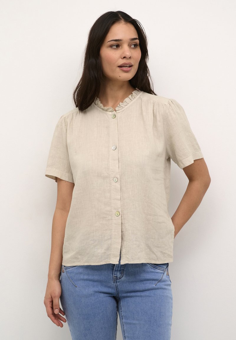 Lys beige kortærmet bluse med flæset krave og knapfront. Lavet af hør, med subtil tekstur og to knapper ved halsen.
