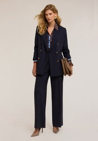 Granatowy żakiet dwurzędowy z złotymi guzikami, zestawiony z szerokimi spodniami i wzorzystą bluzką. Dodatki to brązowa torebka i szpilki.