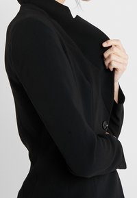 Zwarte op maat gemaakte blazer met een hoge halslijn, gestructureerde schouders en een enkele knoopsluiting. Soepele stof met een aansluitende pasvorm.