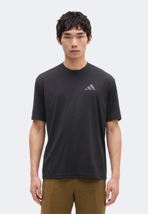 Ung mand iført en sort kortærmet Adidas T-shirt med logo på brystet og brune bukser, står mod en ensartet lys baggrund.