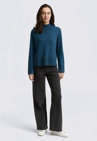 Kék kötött pulóver magas nyakkal, laza szabással és bordázott szegéllyel, fekete bőszárú farmerrel és fehér sportcipővel párosítva.