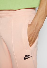 Světle růžové tepláky z měkkého materiálu se bočními kapsami, s černým logem Nike na levé stehno, nošeny s zeleným tričkem.