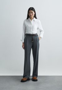 Bijela košulja dugih rukava s diskretnim nabranim detaljima, uparena s hlačama visokog struka u sivoj boji, koje imaju smeđi kožni pojas i debelim crnim cipelama.