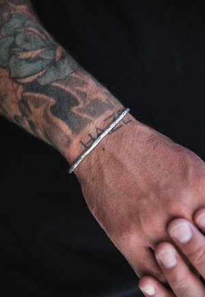 Silver bracelet with a twisted design worn on a tattooed wrist, featuring a textured surface and a minimalist, unadorned style.