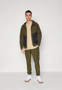 Homme debout portant une veste vert olive avec des panneaux noirs, un pantalon cargo assorti, une chemise beige et des baskets blanches sur un fond uni.