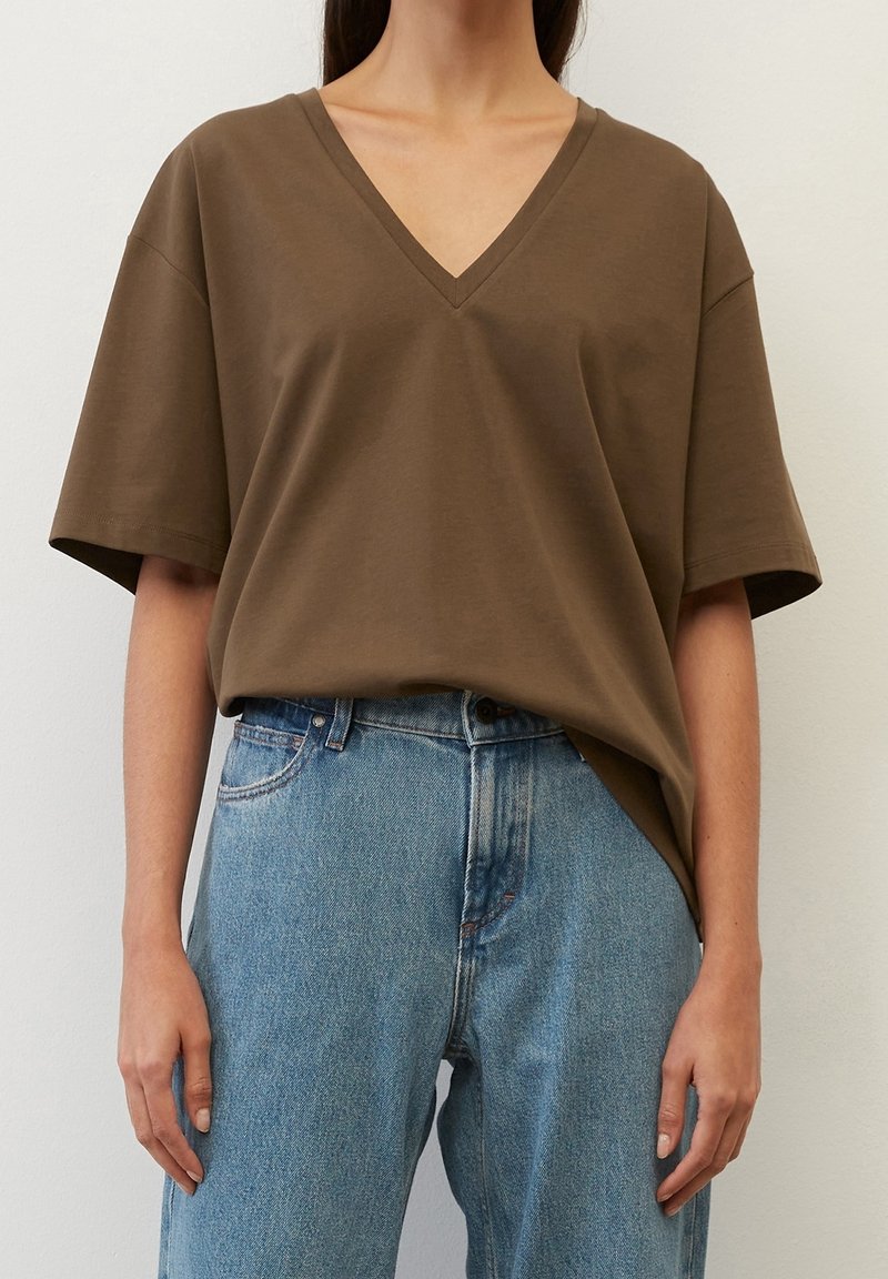 Personne portant un t-shirt marron à manches courtes avec col en V, rentré dans un jean bleu taille haute, sur un fond uni.
