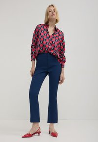 Frau mit blonden Haaren, die eine rot-blau gemusterte Bluse, eng anliegende marineblaue Hose und rote Slingback-Schuhe mit Kitten-Heels trägt, steht vor einem schlichten Hintergrund.