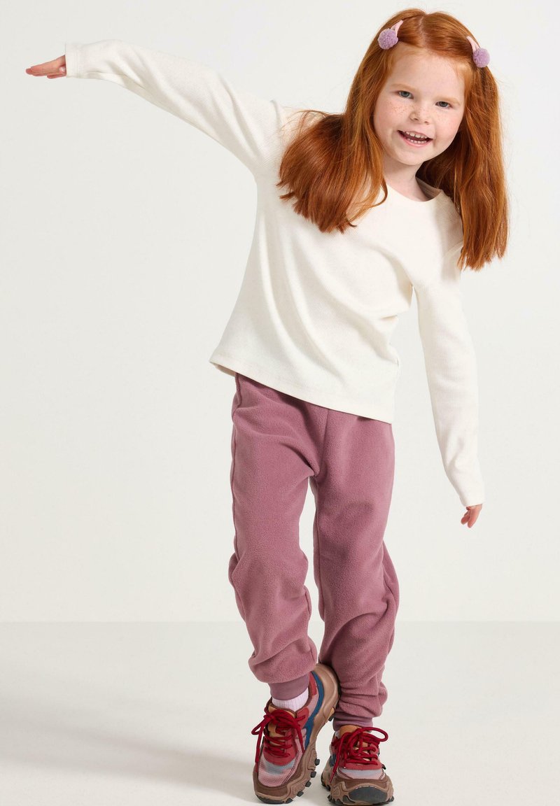 Weißes Langarmshirt mit weicher Rippenstruktur, kombiniert mit rosa Fleecehosen. Sneakers in mehreren Farben mit roten Schnürsenkeln. Kind in verspielter Pose.
