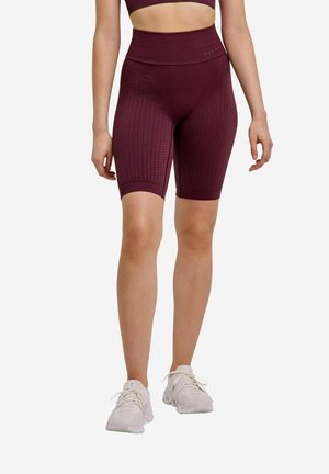 Mujer con pantalones cortos de ciclista de talle alto y textura granate y zapatillas deportivas blancas, de pie frente a un fondo liso.