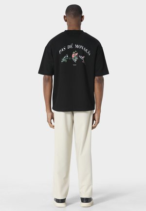 Mand iført en sort oversized T-shirt med "PAS DÉ MONACO" og blomsterbroderi på ryggen, cremefarvede bukser og sorte sneakers.