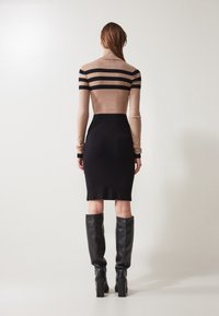 Donna che indossa un vestito a collo alto a strisce beige e nere con una gonna nera e stivali neri fino al ginocchio, in piedi di spalle su uno sfondo uniforme.