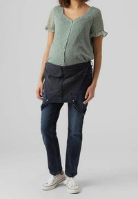 Hellgrüne Bluse mit kurzen Ärmeln und Polka-Dots, vorne mit Knöpfen; verstellbare, dunkelblaue Jeansschürze mit Taschen; schmal geschnittene dunkle Jeans; weiße Sneakers.