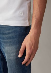 Weißes Baumwoll-T-Shirt mit einem subtilen, strukturierten Logo am Saum, kombiniert mit blauen Denim-Jeans, die eine verblasste Optik und leichte Waschungen aufweisen.