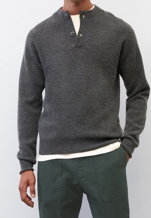 Mann trägt einen dunkelgrauen gestrickten Pullover mit geknöpftem Kragen und dunkelgrünen Hosen, steht mit einer Hand in der Tasche.