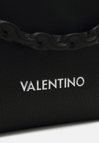 Melnas ādas soma ar viļņotu malu, uz kuras priekšējās daļas ir sudraba "VALENTINO" logotips. Teksturējums piešķir vizuālu interesi.