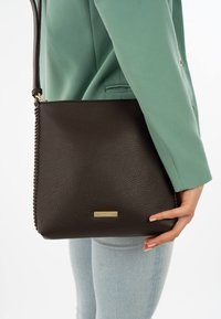 Brown leather crossbody bag with a textured finish, featuring a braided detail along the side and a small gold-tone logo plate.