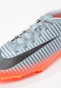 Šedé a oranžové fotbalové kopačky s texturovaným svrškem s vertikálními pruhy, černým logem Nike a oranžovými trny pro trakci.