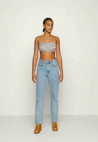 Mulher jovem com cabelo trançado, vestindo um top curto às riscas, jeans clarinhos de cintura alta e botas de cano curto castanhas de salto, de pé contra um fundo simples.