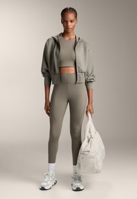 Športna obleka v umirjeni zeleni barvi, ki vključuje krojeno kapuco in usklajene legice z detajli na panelih. Bele superge in lažna torba.