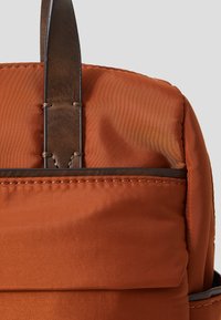 Zaino in nylon marrone con finitura testurizzata, dotato di spallacci in pelle e dettagli di cucitura. Visibile la parte superiore arrotondata e una tasca frontale.