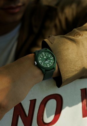 Montre-bracelet verte avec un bracelet texturé, arborant de grands chiffres blancs sur un cadran vert assorti, posée sur une manche et un panneau marron.