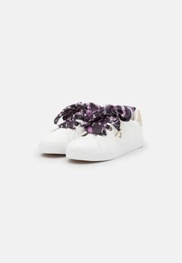 Sneakers sintetici bianchi con una finitura testurizzata, grandi fiocchi a motivi viola e neri come lacci, e un accento con logo dorato.