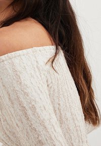 Kvinna med långt mörkt hår iklädd en texturerad crèmefärgad off-shoulder-tröja, som visar bar axel och överarm.