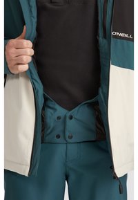 Chaqueta aislante en tonos verde azulado y negro, con cierre delantero con cremallera y botones a presión, forrada interiormente con material suave y puños elásticos.