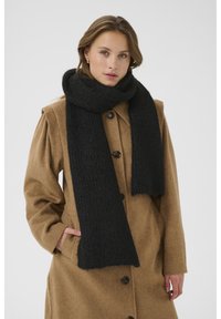 Musthave mustra kääritud musta kudumiskaeluse ja tekstuuriga mustriga, kontrastniveerituna beeži villase jopega, millel on suured nööbid ja taskud.