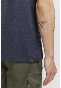 Temno modra kratka majica s teksturiranim zaključkom, s rjavim etiketom na robu in zelenimi vojaškimi hlačami z stranskim žepom.