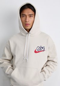 En ung mand med langt hår iført en beige hoodie med et legende logo. Han står foran en ensfarvet baggrund og ser direkte på beskueren.