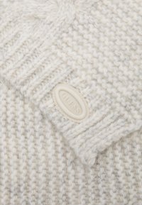 Maglione bianco a maglia con un motivo testurizzato. Presenta un colletto cucito e un'etichetta bianca sporca con la scritta "GUESS," situata di lato.
